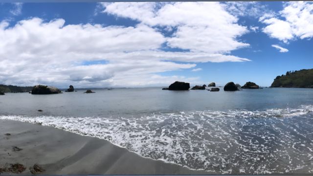 Surf running up a beach toward sea stacks under a cloudy sky